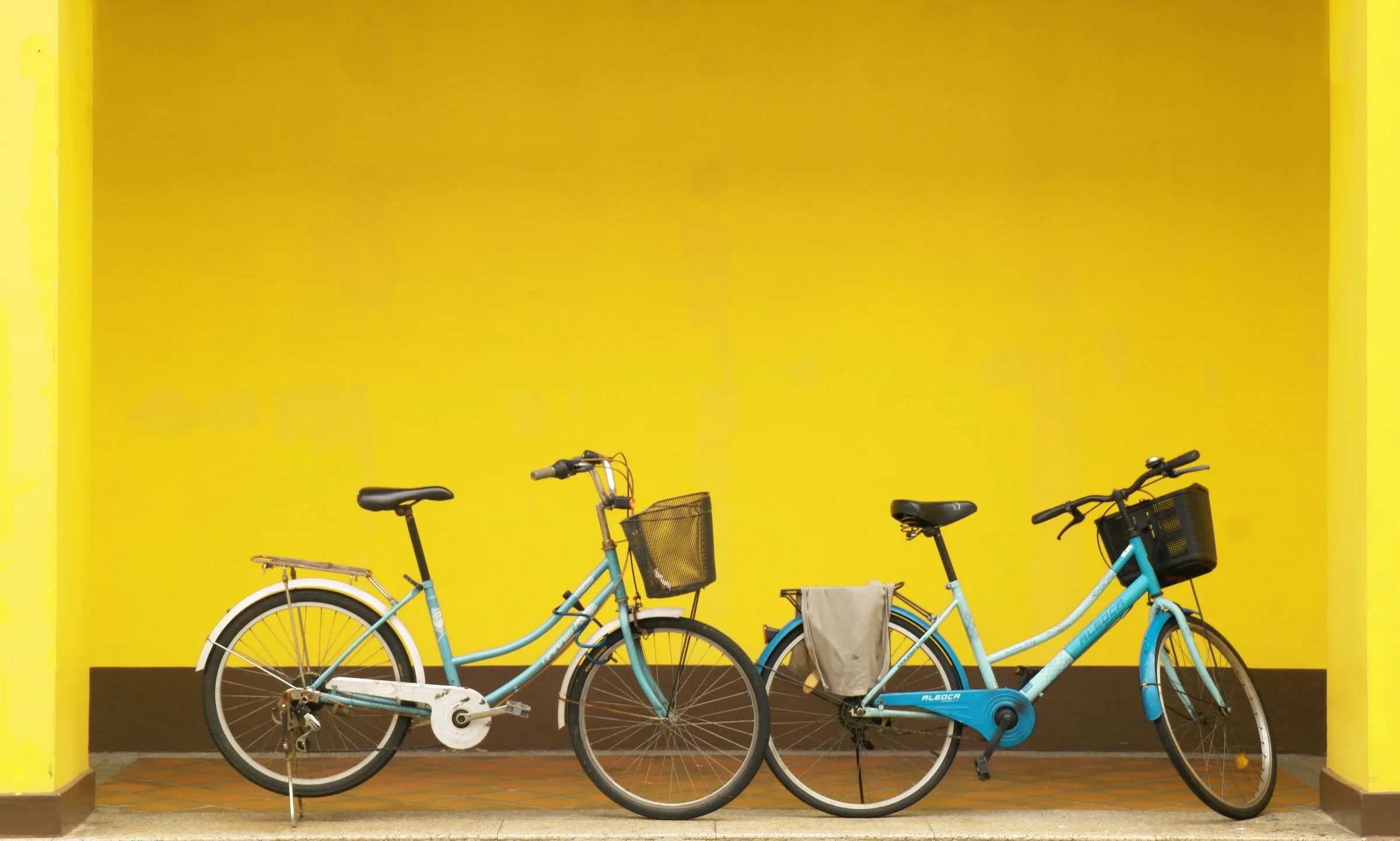 Two city bikes with baskets in front of a yellow wall